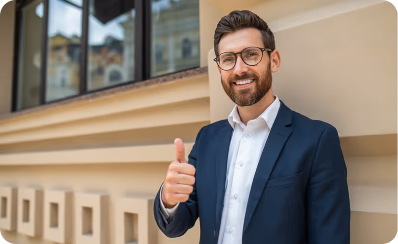 Smiling man wearing glasses and a dark suit giving a thumbs-up in front of a beige building.