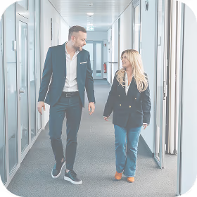 A casually dressed man and woman walking and talking in a modern office hallway.