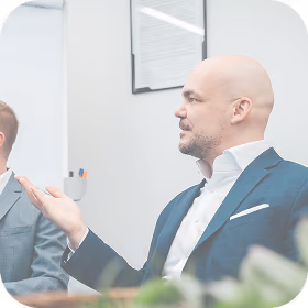 Bald man in a blue suit gestures while speaking in an office setting.