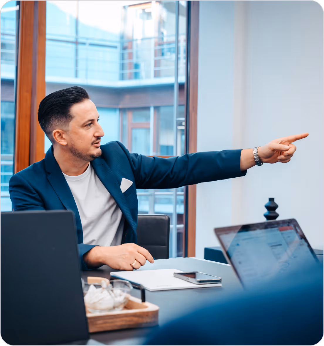 Man in a blue blazer pointing to something off-camera during a meeting at a modern office table with laptops and a smartphone.