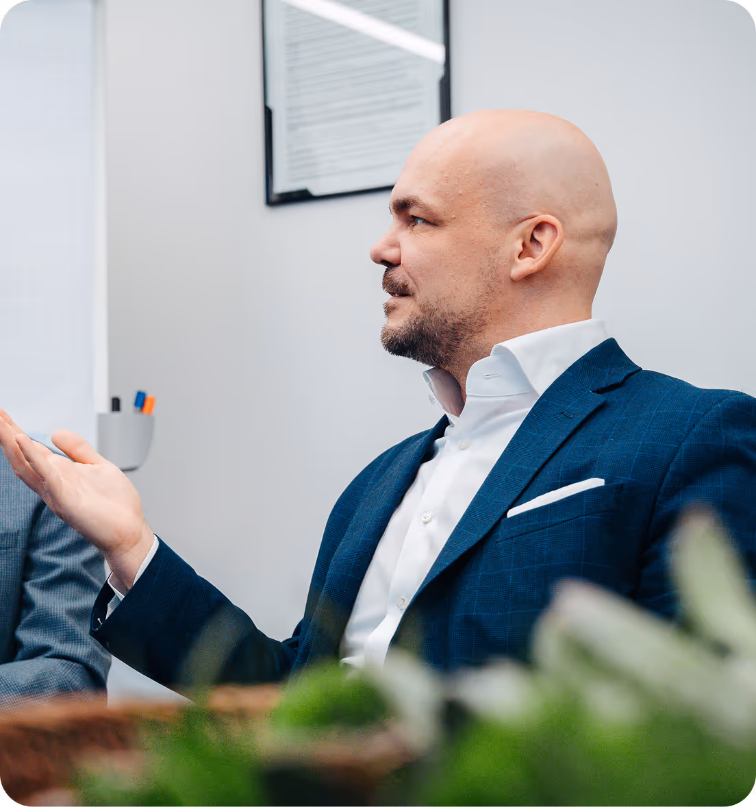 Bald man in a blue suit and white shirt gesturing with his hand during a conversation in an office.