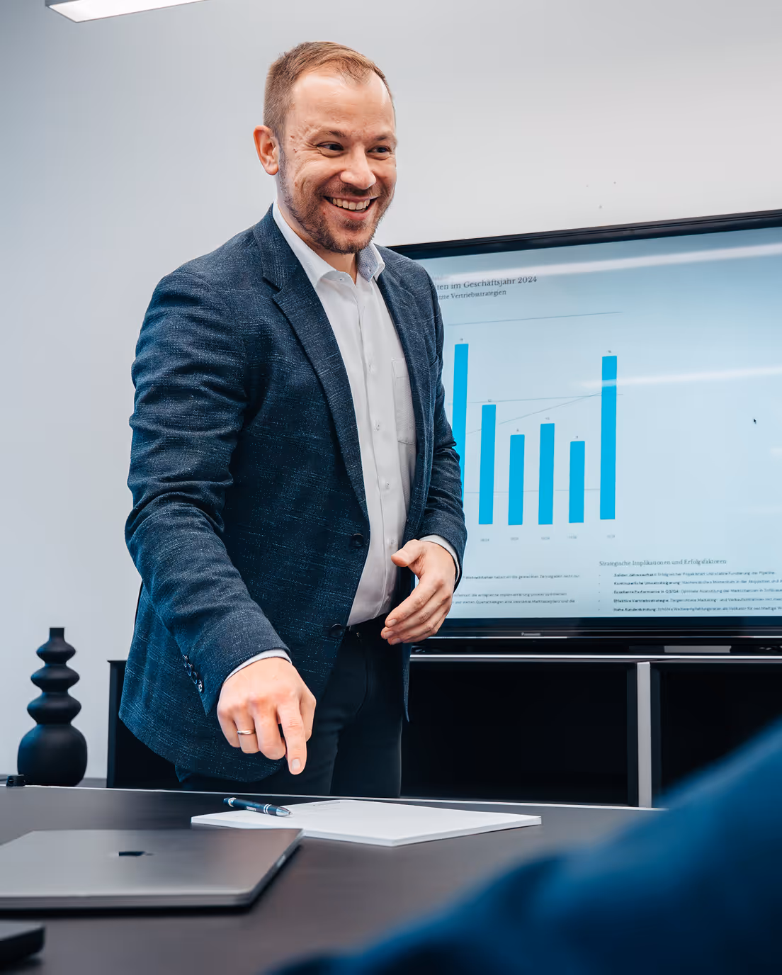 Smiling man in business attire pointing at a notebook on a table with a laptop, with a presentation screen showing blue bar charts in the background.