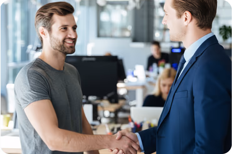 Two men shaking hands in an office, one wearing a gray t-shirt and the other a blue suit.