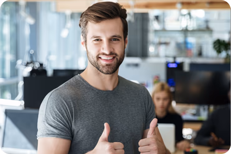 Smiling man in gray t-shirt giving two thumbs up in a modern office setting.
