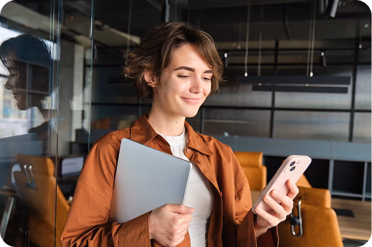 Junge Frau im braunen Hemd hält einen Laptop und schaut lächelnd auf ihr Smartphone in einem modernen Büro.