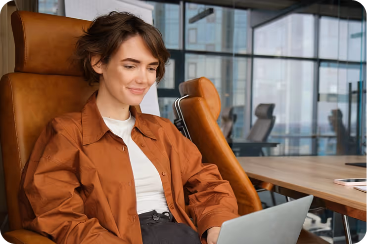 Junge Frau in brauner Jacke arbeitet am Laptop in einem modernen Büro mit großen Fenstern.