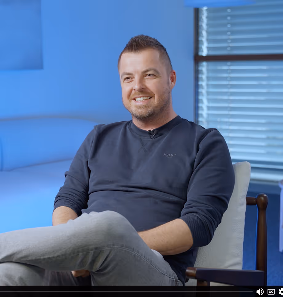 Ein lächelnder Mann in dunkelblauem Pullover sitzt entspannt auf einem Stuhl vor einem Fenster mit Jalousien.