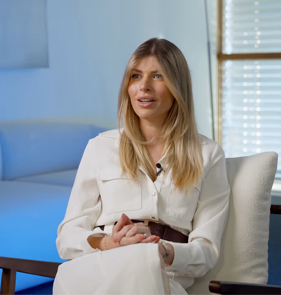 Frau mit langen blonden Haaren, die in einem hellen Raum auf einem Stuhl sitzt und spricht.