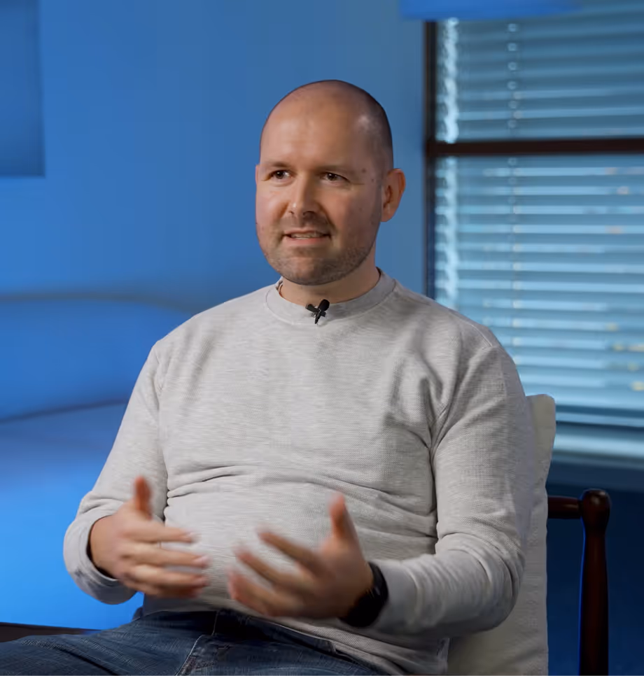 Ein Mann mit grauem Sweatshirt sitzt vor blauem Hintergrund und spricht gestikulierend.