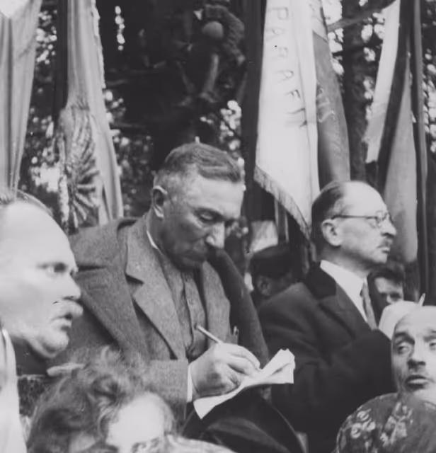 Black and white photo of a man in a suit writing on paper, surrounded by several other men with flags in the background.