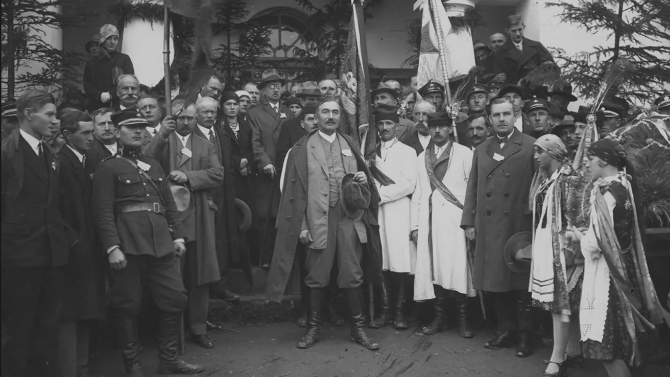 Black and white historical group photo of men in formal and military attire and women in traditional dresses, standing outside a building.