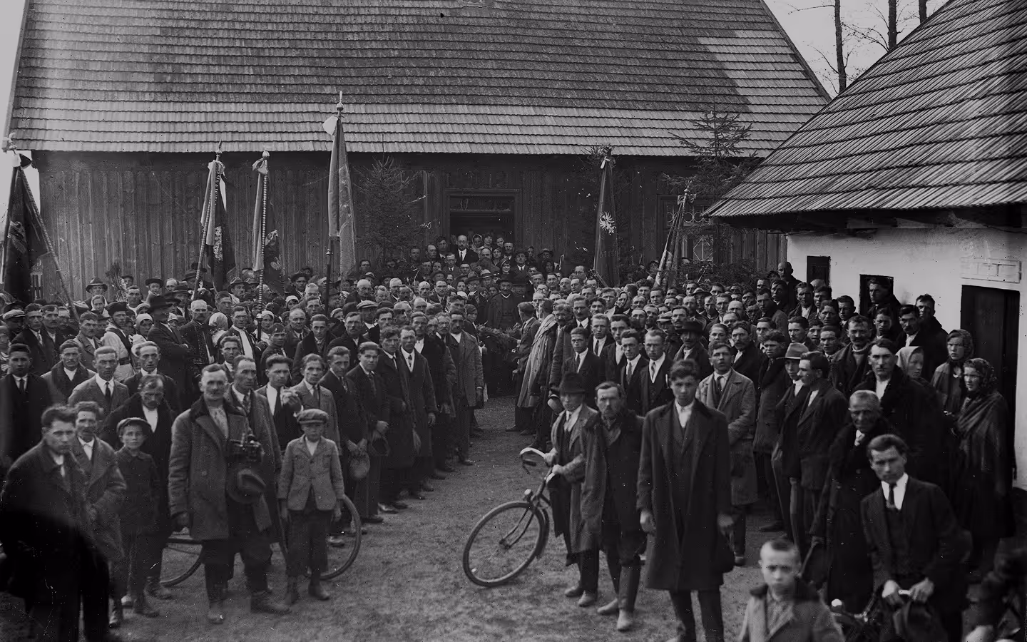 Large black-and-white group photo of men, mostly in suits and coats, standing outdoors between two wooden buildings, with some holding flags.