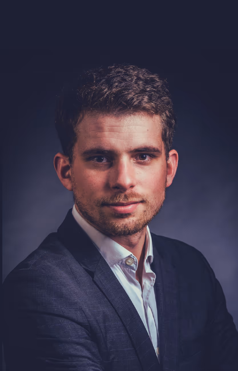 Portrait of a confident young man with short curly hair, wearing a dark blazer and white shirt against a dark background.
