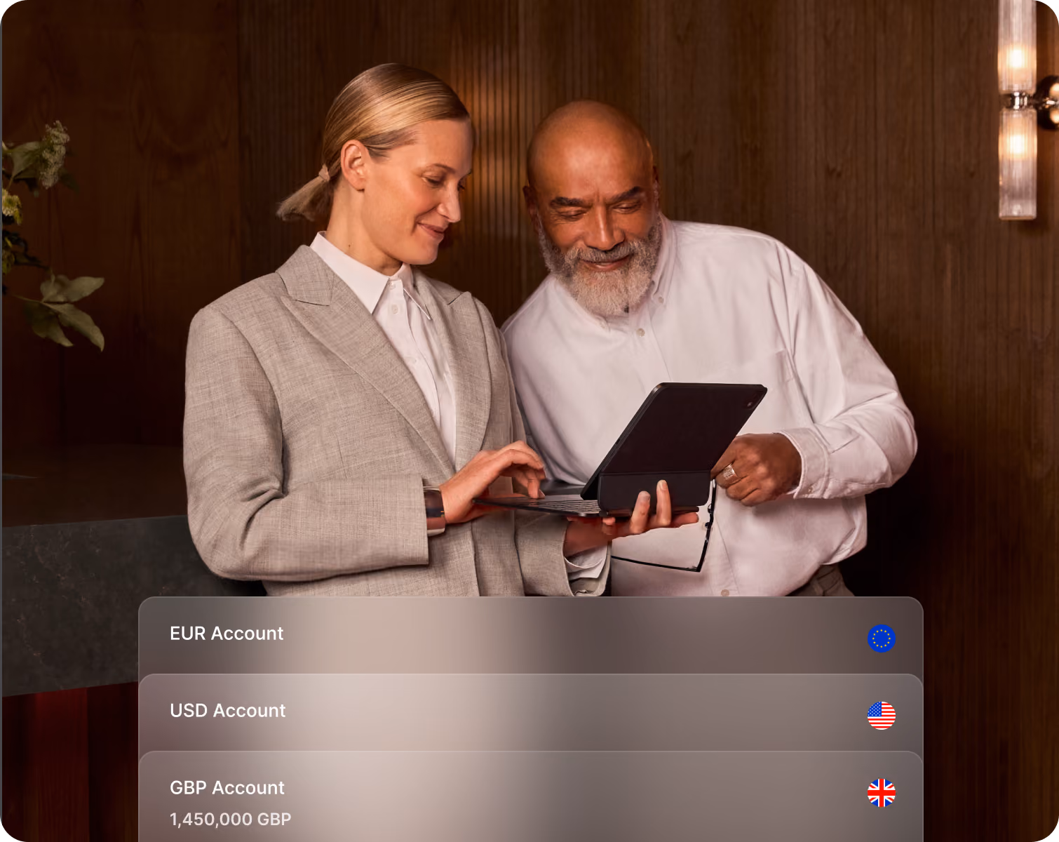Two people smiling and viewing a tablet, with a currency account list overlay shown on screen.