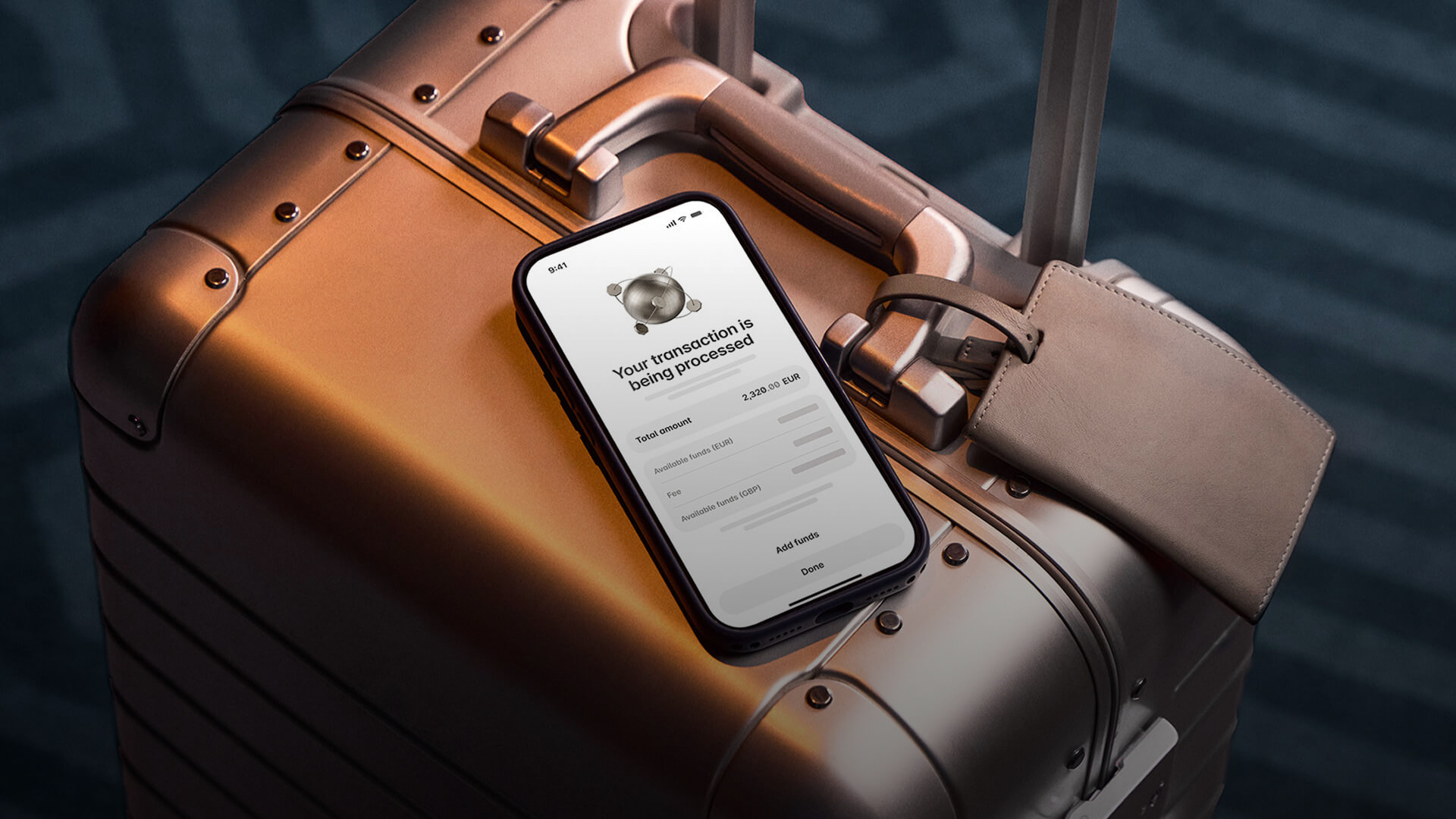 Smartphone showing a "transaction processing" screen for a 2,320.00 EUR transfer, resting on a bronze hard-shell suitcase with a leather luggage tag.