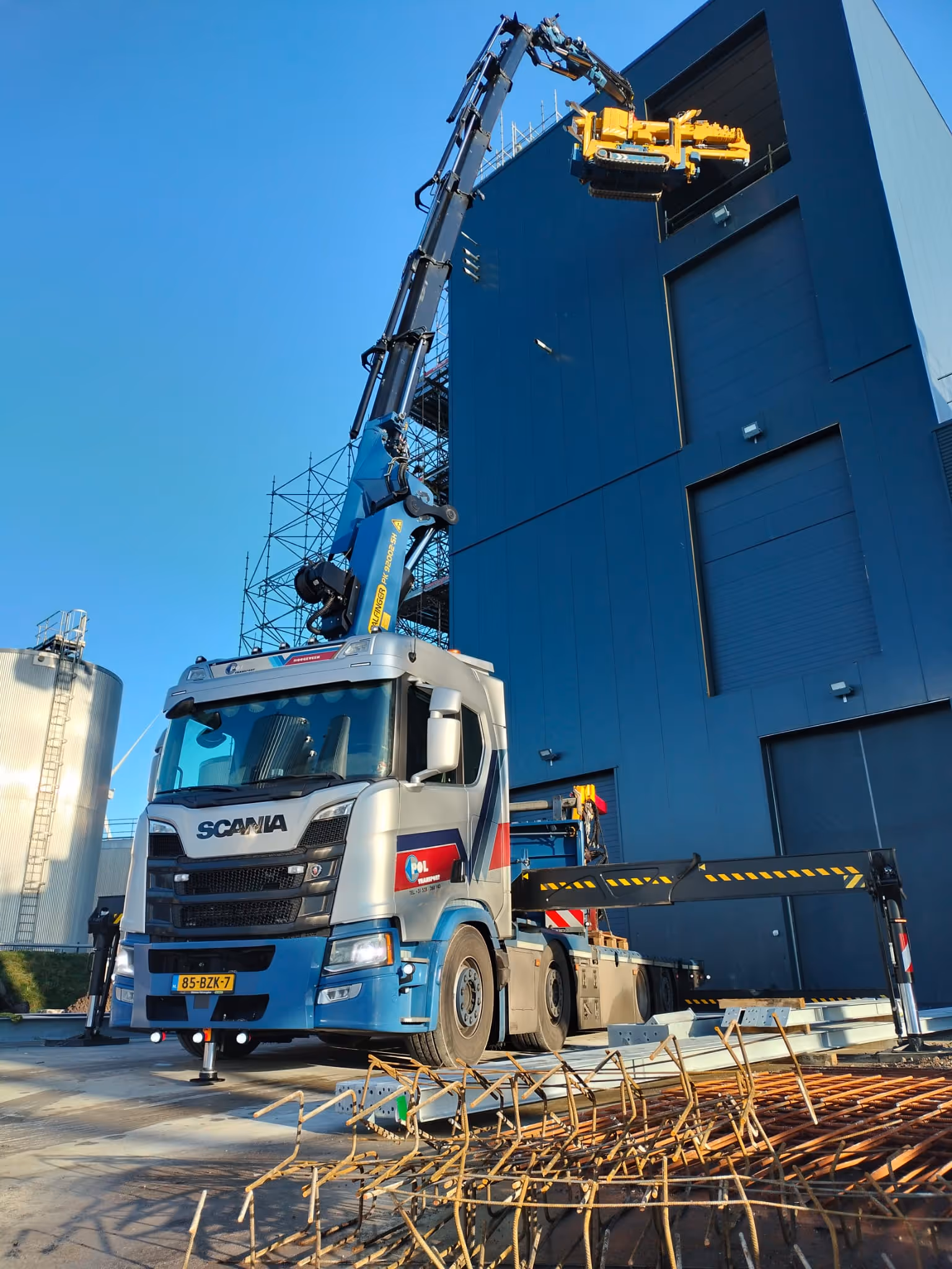 Blauwe en zilveren Scania vrachtwagen met opbouwkraan die een gele machine omhoog tilt bij een grote blauwe hal.