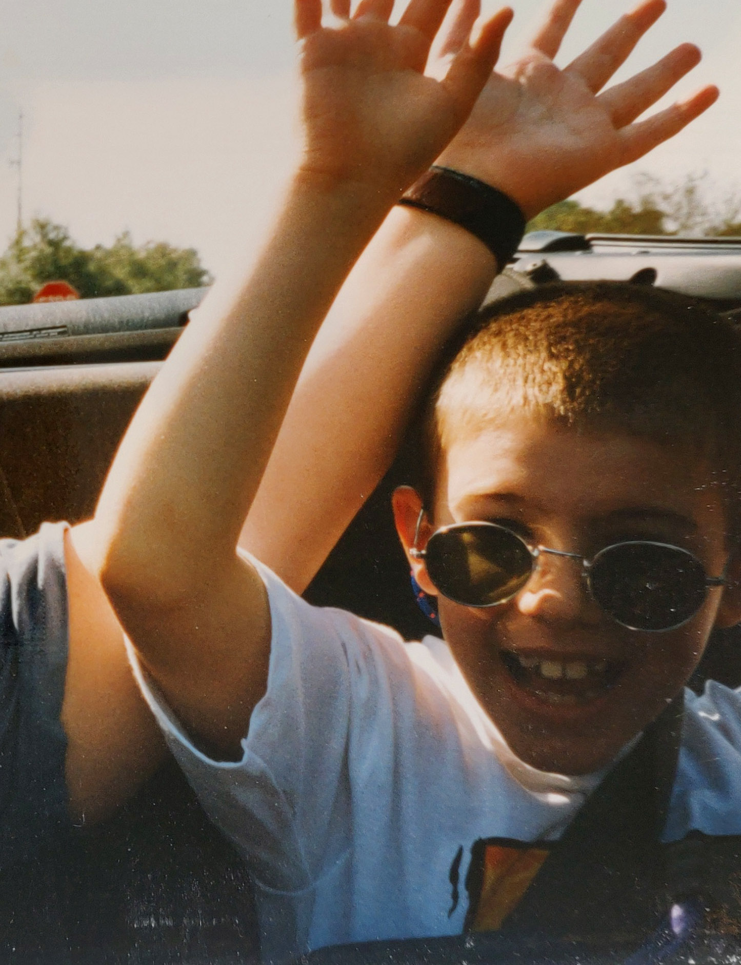A smiling young boy wearing round sunglasses and a white shirt raises his hands up, sitting in a car with another person whose hands are also raised.