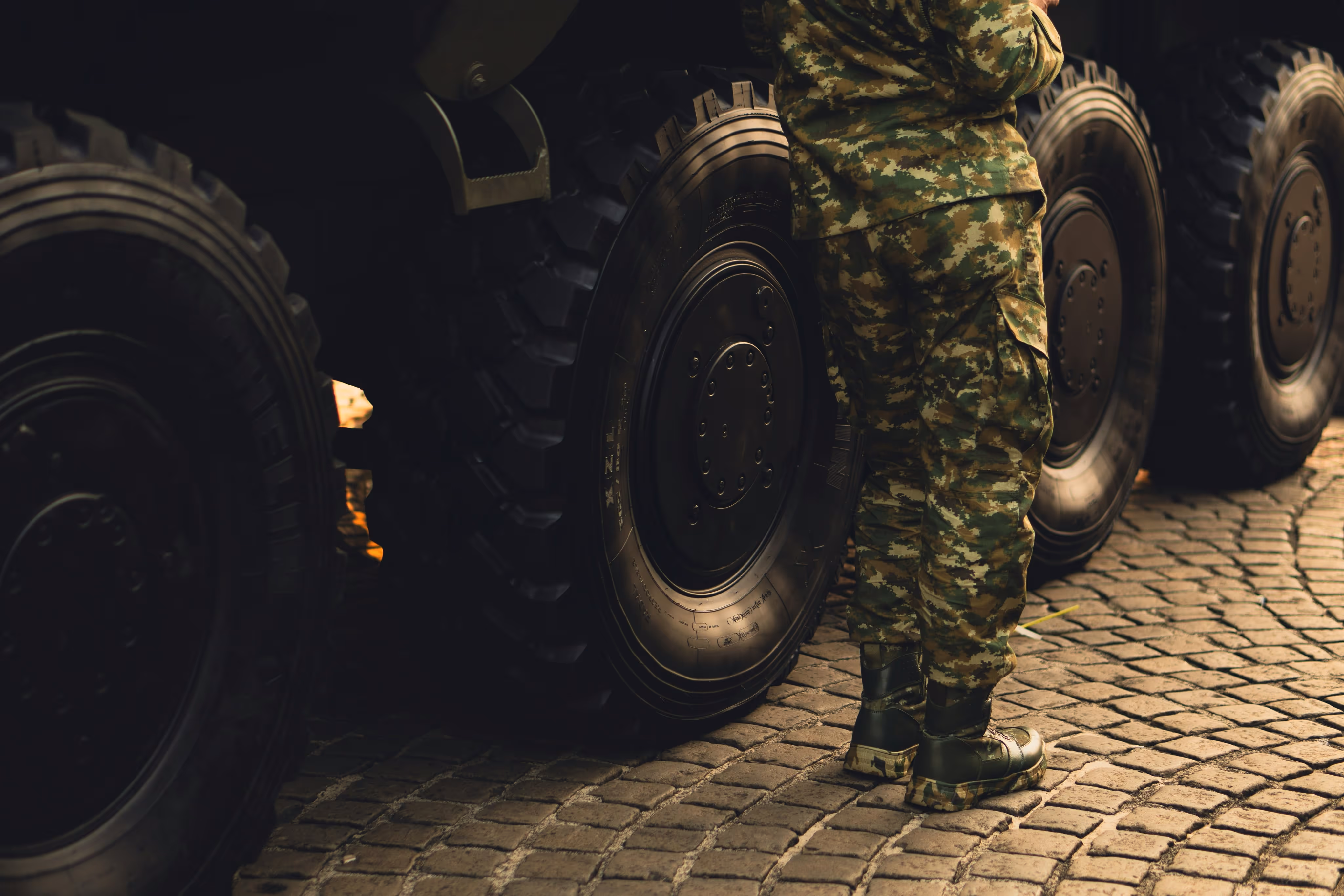Person wearing military camouflage uniform and boots standing beside large armored vehicle tires on cobblestone ground.