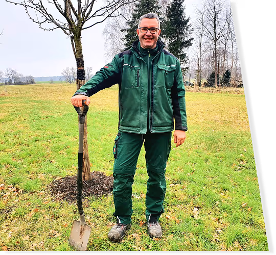 Mann in grüner Arbeitskleidung steht auf einer Wiese und lehnt sich an eine Spatschaufel.