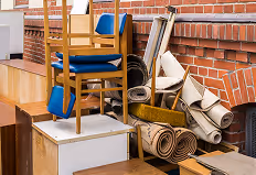 Gestapelte Möbelstücke, darunter ein Holzstuhl mit blauem Sitz, weiße Kisten und zusammengerollte Teppiche vor einer Ziegelwand.