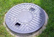 Runder Metallkanaldeckel auf Gras mit rechteckigen Grifföffnungen.