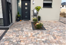 Haustür mit grauen gepflasterten Gehweg und bepflanzten Blumenbeeten vor einem modernen Haus.