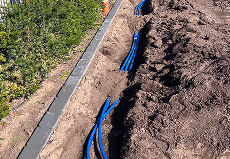 Blaue Kabel oder Rohre, die in einem gegrabenen Graben neben einem Gehweg mit Pflanzen verlegt werden.