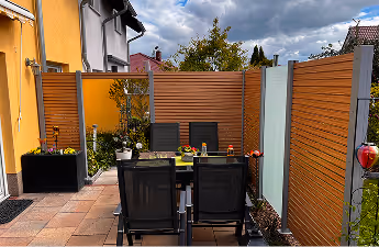 Schattiger Terrassenbereich mit schwarzem Gartentisch und vier Stühlen, von Holzsichtschutzwänden umgeben und vor gelber Hauswand.