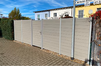 Moderner grauer Sichtschutzzaun mit einer Tür entlang eines gepflasterten Gehwegs vor Wohngebäuden.