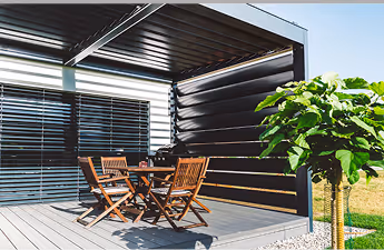 Moderner Außenbereich mit Holztisch und Stühlen unter einer Pergola mit Lamellen, daneben ein großer grüner Baum.