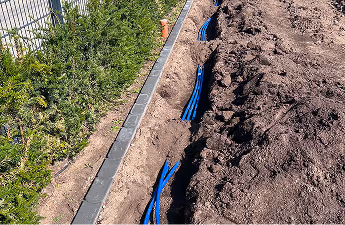 Im Boden verlegte blaue Rohrleitungen neben einem Pflasterweg und einer Wiese, mit aufgeschüttetem Erdreich rechts.