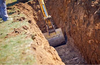 Bagger hebt Erde aus einem tiefen Graben, großer Erdwall links, Person steht am Grabenrand.