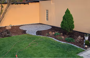 Ecke eines Gartens mit grünem Rasen, einem gepflasterten Rundweg, Büschen und einem Baum vor einer gelben Hauswand.