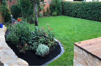 Gepflegter Garten mit grünem Rasen, Hecke im Hintergrund und einem Bereich mit Kräutern und Pflanzen neben einem gepflasterten Weg.