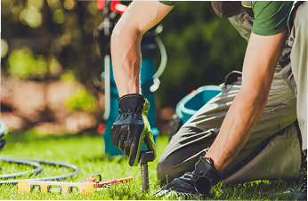 Person kniet auf Gras und benutzt ein Werkzeug, wahrscheinlich zum Gartenarbeiten oder Pflanzen.