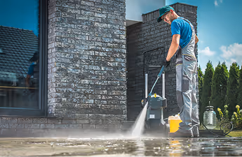 Mann reinigt mit Hochdruckreiniger eine gepflasterte Fläche vor einem modernen Haus.