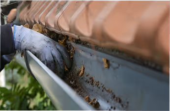 Hand in Arbeitshandschuh reinigt mit einem kleinen Werkzeug das Dachrinnensystem, entfernt Blätter und Schmutz.