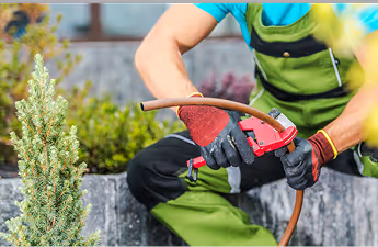 Person in grüner Arbeitskleidung und Handschuhen repariert ein Gartenschlauch mit einem Werkzeug vor einer Steinmauer.