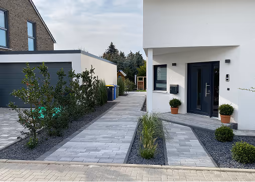 Moderner Hauseingang mit weißer Fassade, grauem Pflasterweg und gepflegten grünen Pflanzen in grauem Kiesbett.