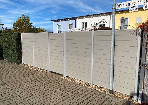 Ein beiger Sichtschutzzaun mit Tor neben einem gepflasterten Gehweg unter blauem Himmel.