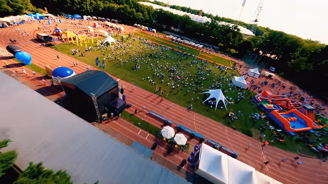 Aerial view of an outdoor event on a sports field with a crowd, tents, inflatable structures, and a stage near a running track.