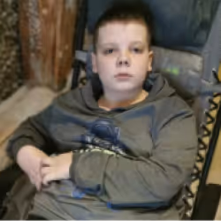 Young boy with short hair wearing a gray hoodie sitting indoors, looking directly at the camera with a neutral expression.