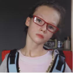 Young person with short hair wearing red glasses and a light pink shirt sitting against a dark background.
