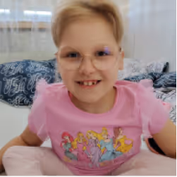 Smiling young child with glasses and short blond hair wearing a pink shirt with a princess design.