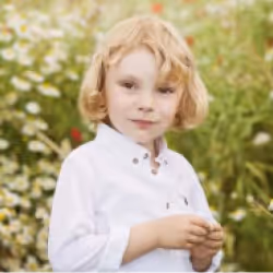 Young child with blonde hair wearing a white shirt standing outdoors with greenery in the background.