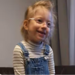 Young child with glasses wearing a striped turtleneck and denim overalls smiling indoors.