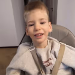 Smiling young boy with light brown hair sitting in a chair with beige straps over his shoulders.