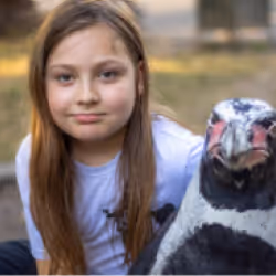 Young girl with long brown hair smiling next to a penguin statue outdoors.