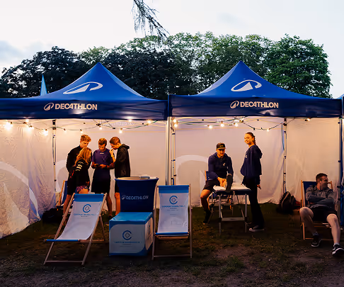 Decathlon branded outdoor booth with tents and deck chairs, people standing and sitting under warm string lights.