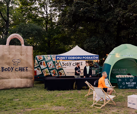 Outdoor event with Body Chief branded inflatable bag, food truck, and tent serving meals with people sitting and standing nearby.